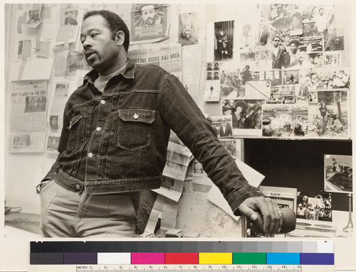 Eldridge Cleaver in front of press clippings on wall of room in Paris