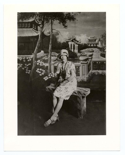 Tress in knee-length, low-necked dress with cloche hat and high-heeled strap shoes sitting on a bench against Chinese park background/