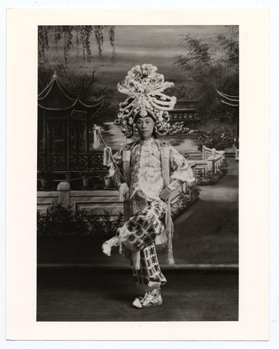 Actor in role of a gallant with an elaborate headdress holds a riding crop with one leg raised /