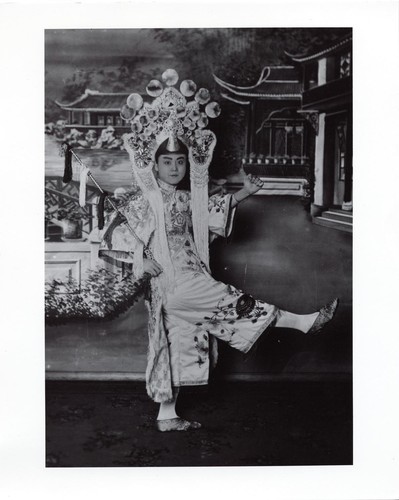 Portrait of an actor in classical costume holding a whip, one leg raised on Chinese garden with pavilions background /