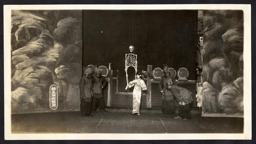 Woman stands among four demon attendants, rows of coffins and skeleton in the background /