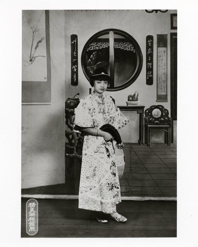 Actress in modern 1920s dress with wide sleeves and "ear muff" hairdo holding a folding fan /