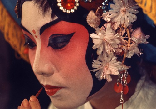 Close-up of a Chinese actress wearing theatrical makeup applying red lipstick and wearing a cap with floral and jewel decorations /