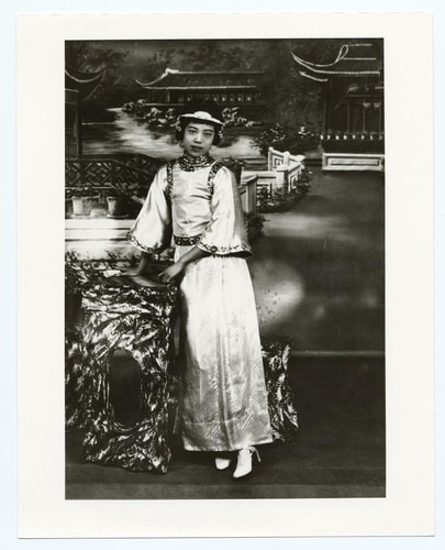 Portrait of an actress with bobbed hair, wide sleeved jacket and ankle length skirt standing by a table /