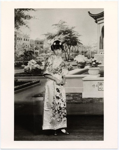 Standing actress in modern dress with wide sleeves and "ear-muff" hairdo with a backdrop of a city and park /