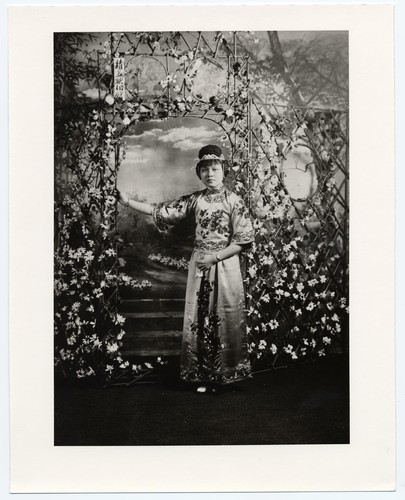 Actress in early 20th century ankle-length skirt and jacket standing in a bamboo arbor /