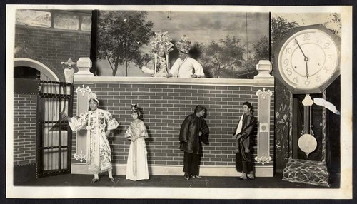 A man points to the left a woman follows, two servants stand by a large clock and two other men observe the scene from above, staged at the Great Star Theatre /