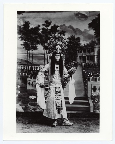 Scholar in full classical costume and elaborate headdress holds a fan and a book /