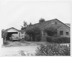 Two people stand next to a Ford coupe outside the attached garage of a single-story brick house in an unidentified Sonoma County, California, location, late 1940s or 1950s