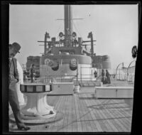 A man peeks at the camera while standing in front of gun turrets aboard a battleship off the coast, Santa Monica, about 1900