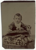 Portrait of an infant in plaid dress and boots on embroidered chair