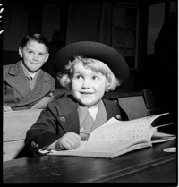 [First French Lesson: small child, Hans, in dark (Alsatian?) suit and round hat, in school room with older children]