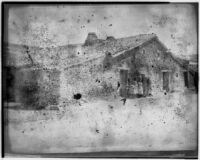 Group of people standing in front of a low stone house with a clay tile roof, Europe, 1929