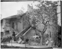 Woman in a floppy hat sitting on the stone railing of a staircase that leads down from a stucco house, Europe, 1929