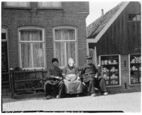 Woman and two men in traditional Dutch clothing sitting in front of a brick building, Volendam, Netherlands, 1929