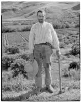 Man holding a straw hat and walking stick, standing on a hill with fields behind him, Europe, 1920s