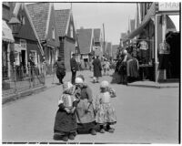 Three little girls wearing traditional Dutch clothing with a crowd of shoppers in the background, Holland, 1929