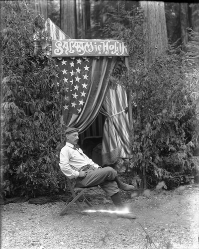 Man, identified as Marcus, sitting in folding chair by tent with sign above door "Salaam ie Hogir," Bohemian Grove. [negative]