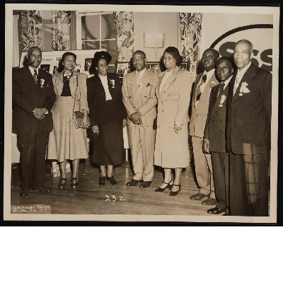 Group photograph of eight men and women standing in front of Brotherhood of Sleeping Car Porter posters