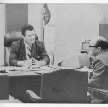 Duane Lowe, seated at his desk, talks with Frank Rogers