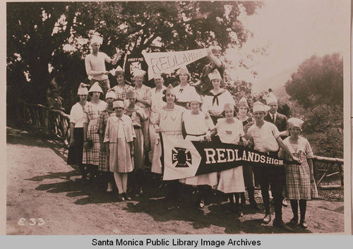 Group of school children with matching hats stand together holding a banner "Redlands High" in front of a large oak tree, Pacific Palisades, Calif