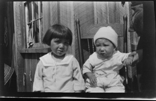 Two Native American children sitting in front of a wooden house