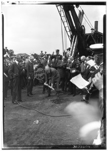 Large crowd of men watching a man break ground at a site, while a large machine and the Los Angeles Police Band appear in the background