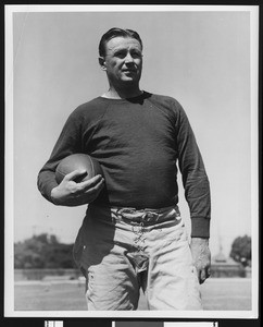 University of Southern California head football coach Jeff Cravath, hands by his sides and holding football, wearing a dark sweatshirt with white t-shirt. Bovard Field. 1942