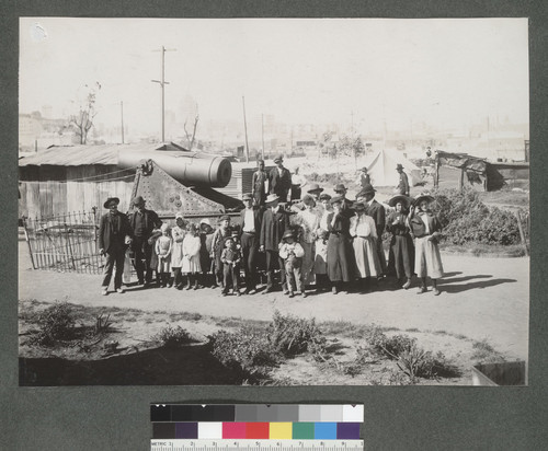 [Group of refugees pose with a damaged cannon. Makeshift shacks and tent in background. Call Building in distance, left center?]