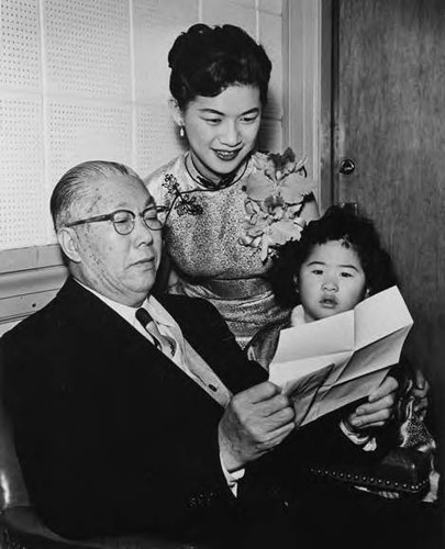 Gee Kee Ward reading a paper with San Francisco's Miss Chinatown wearing a cheong-sam and orchid corsage reading over his shoulder Patricia Gee, his oldest grandchild; possibly on the same occasion as the meeting with the politician, taken in Los Angeles