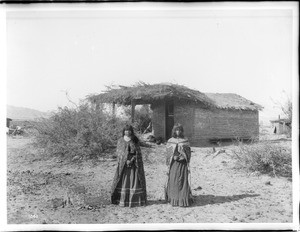 Two Mojave Indians girls standing in front of a small dwelling with a thatched roof, 1900