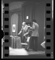 Mickey Cohen is helped into his seat at a question and answer session at the Sheraton Airport Hotel, 1975