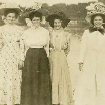 Six women with hats and dresses by the riverbank