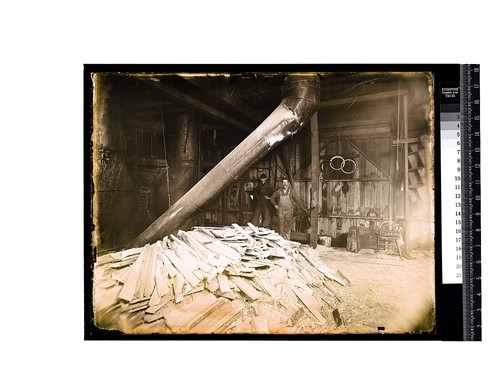 [Two men pose inside a shop near a pile of scrap wood]