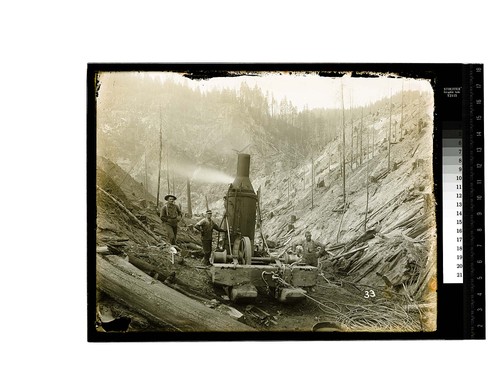 {Three workers pose aroung a steam donkey in logged over hills]