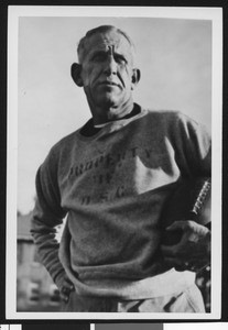 University of Southern California football coach Howard Jones wearing "Property of USC" sweatshirt, holding football, Bovard Field, mid 1930s