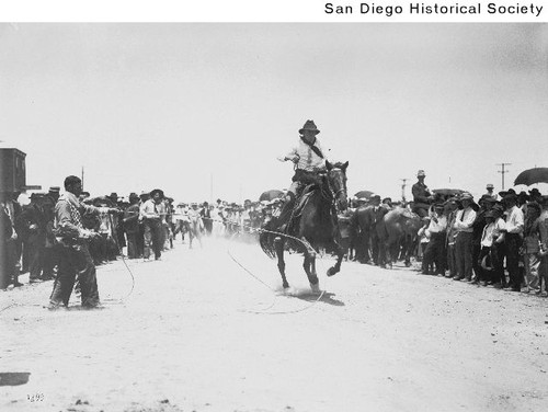Crowd watching a man on a horse riding past a man with a lariat during a cowboy roping exhibition