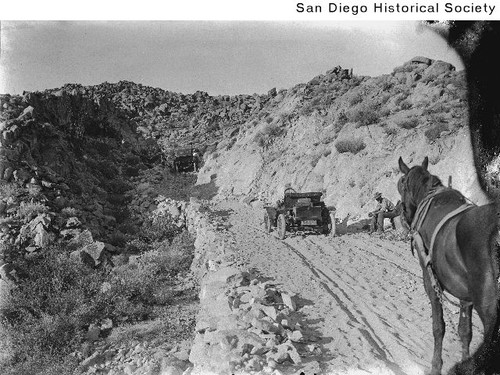 Two automobiles and a horse on an unpaved mountain road with a man sitting by the side of the road