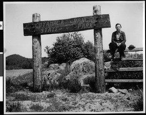 Woman sitting next to the entrance sign to Cuyamaca State Park, ca.1932