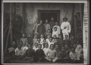 Elementarschule Hoschuwan auf einem Spaziergang vor dem Haus eines Aeltesten in Wongfa. Damals (1909) war die Schule noch klein, heute hat sie 60 Schüler. Sie hatten damals noch Zöpfe & den Vorderkopf rasiert. Miss. Kaufmann