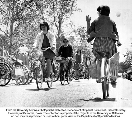 Bicycles, Students riding bicycles through bicycle parking area. Most of the bicycles have baskets. All of the females are wearing dresses or skirts
