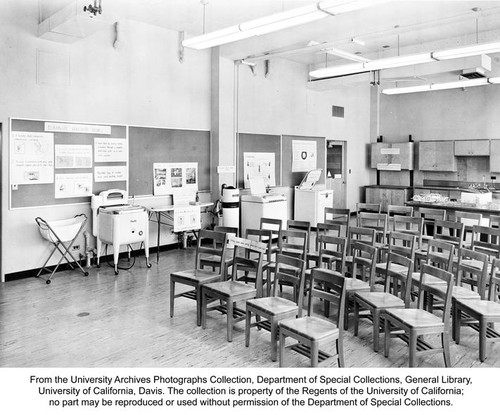 Everson Hall, Home Management Laboratory, Home Economics Building. The Home Management Laboratory in the new Home Economics Building on the Davis campus of the University of California. It is used for both teaching and research in the efficient use of household equipment to reduce fatigue. Modern appliances, designed for function and efficiency, are tested and evaluated. The chairs are of different heights so that students can learn which are best suited to various household tasks. Cabinets, mounted on gears, can be raised and lowered to study proper working heights as an aid in reducing fatigue. Students learn to use cabinet space to best advantage in storing kitchen supplies, with studies of shelf spacings and arrangements. On the table in the rear are various automatic dishwasher racks. These are evaluated for holding capacity and handling ease. Washers are studied for efficiency under home conditions of water temperatures and pressures. A laundry cart, conventional washing machine and ironing board with adjustable legs for height, water-softener, automatic clothes washer, and automatic dishwasher are along the wall