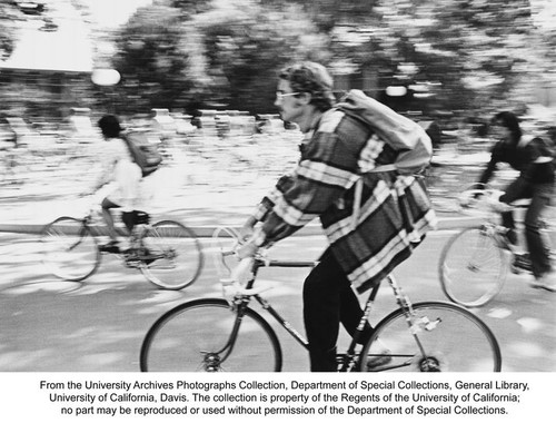 Bicycles, Man riding bicycle wearing plaid jacket, pack on his back. Two other riders in the middle ground and background blurred by motion