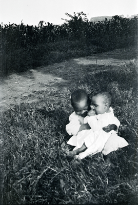Two children sitting on grass, corn field in the background
