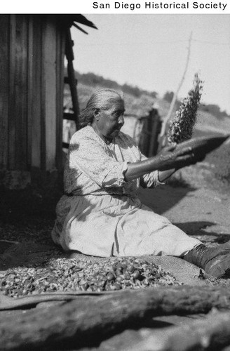 Delores Renterias sitting on the ground outside a house on Palomar mountain, winnowing acorn meal