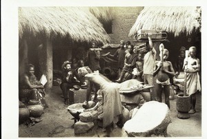 African compound with women grinding maize, girls pounding fufu, carriers for the hammock and the loads. The white man sits on a local stool and in front you see his plate and the calebash for his water. On the left big water pots