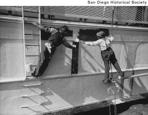 Two men painting the side of a ship