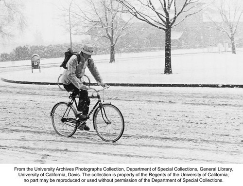 Bicycles, Man wearing jacket, hat, and pack, riding bicycle on slushy California Avenue during snowstorm. The Silo in the background
