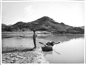 Man standing on shore next to a small ferry boat moored along the Colorado River in one of the Mojave canyons, California, 1900-1950