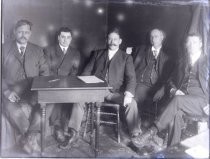 Five men in suits at a table with pen and paper
